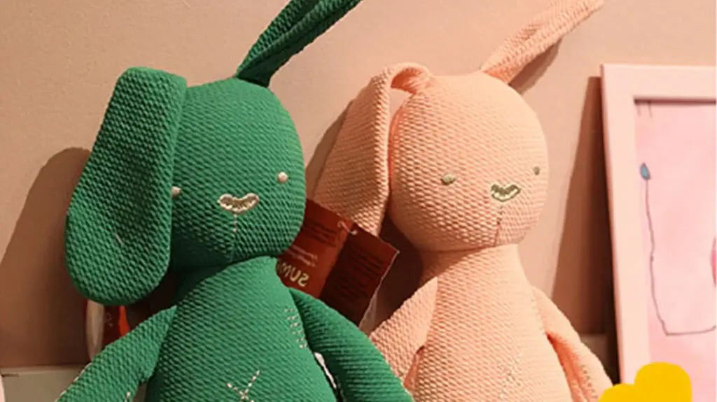 Two soft knitted bunny plush toys, one green and one peach, with simple embroidered faces, leaning against a wall beside a pink picture frame.