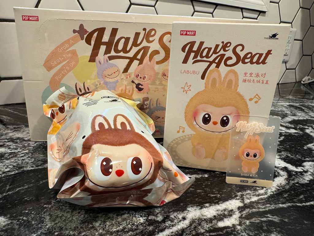 POP MART Labubu “Have A Seat” collectible figure with its box, sealed blind bag packaging, and character card displayed on a marble countertop.