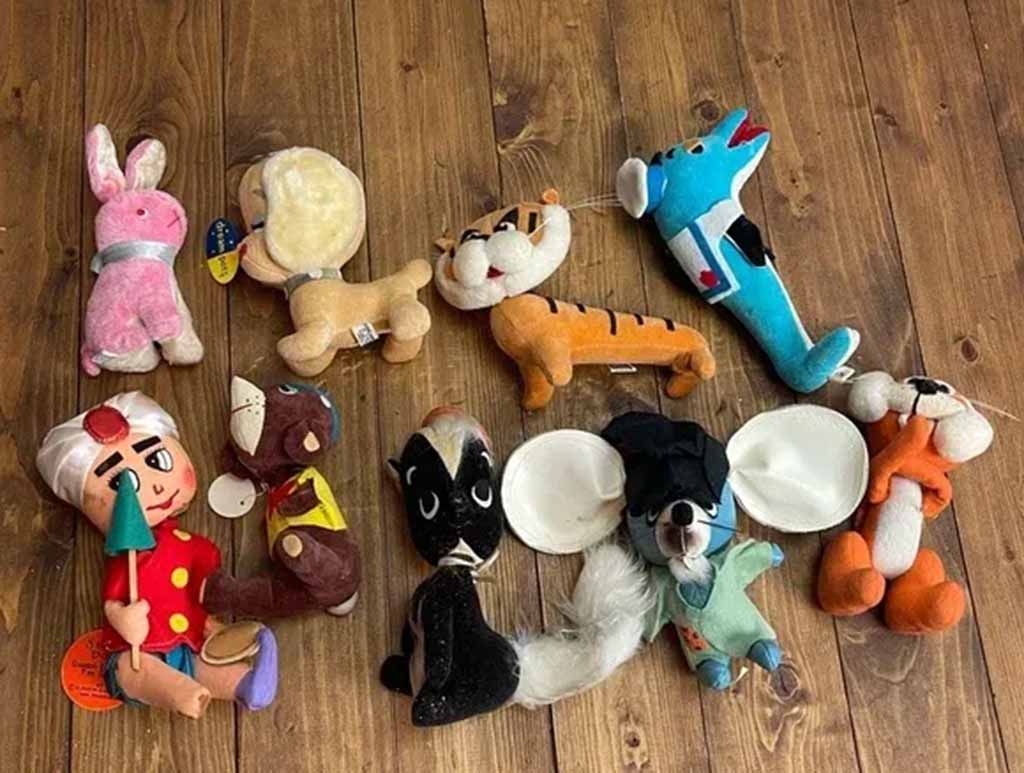 A collection of assorted vintage plush toys arranged on a wooden floor, including animals, cartoon characters, and worn stuffed figures in various colors and styles.