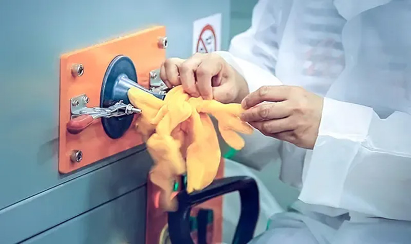 A factory worker uses a stuffing machine to fill a yellow plush toy, with both hands guiding the fabric around the machine nozzle.