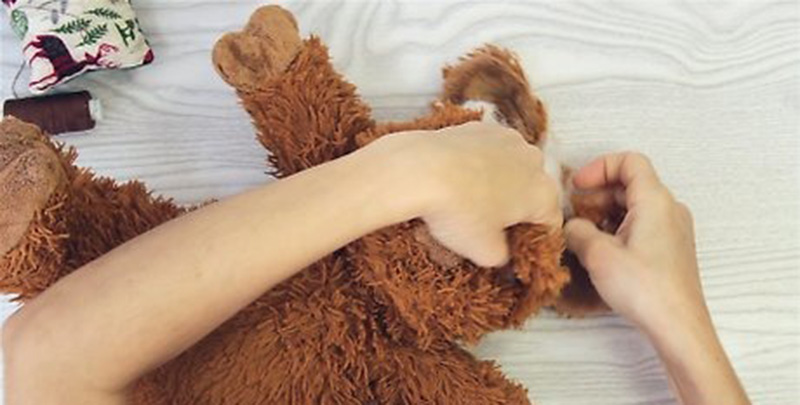 Hands repairing a brown plush toy by opening the seam to adjust or replace internal stuffing on a work surface.