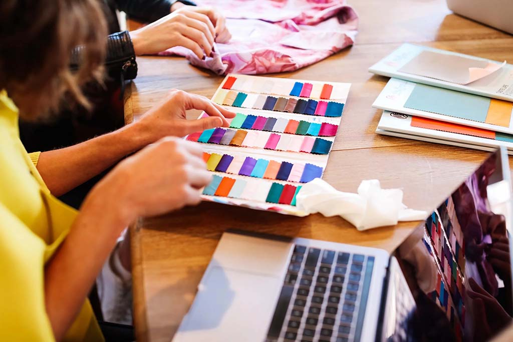 Two people reviewing a fabric color swatch book at a table, comparing textile samples for product design, with a laptop and notebooks nearby during a material selection discussion.