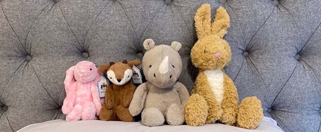 Four plush toys including a bunny, fox, rhino, and rabbit sitting on a bed with a tufted grey headboard in the background.