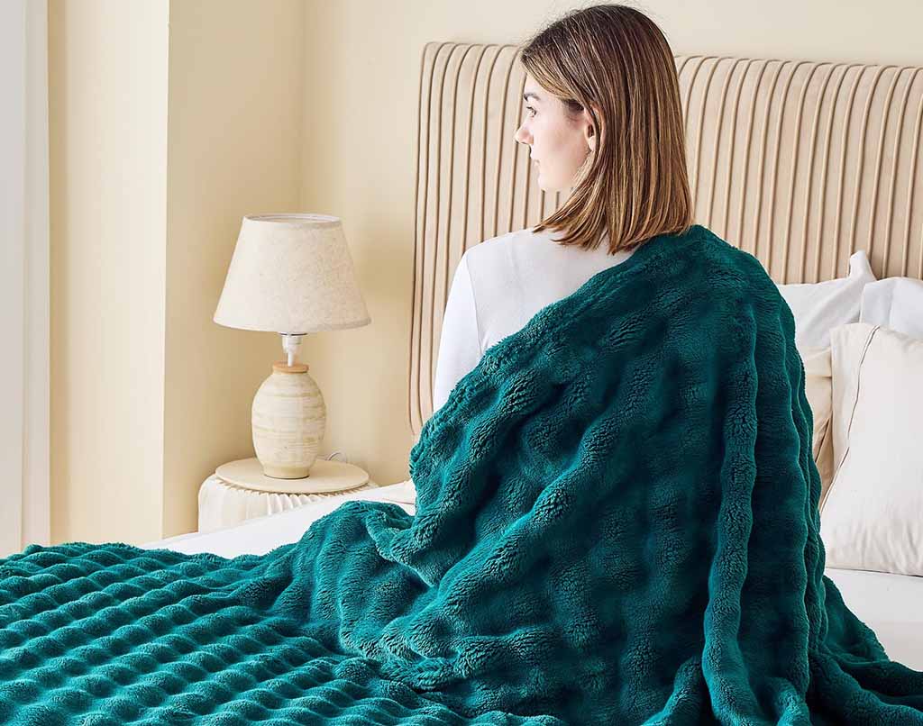 A woman sitting on her bed with a teal textured plush blanket draped over her shoulders, facing away from the camera in a softly lit bedroom.