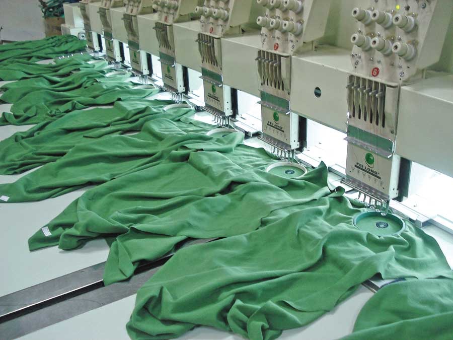 Multiple automated embroidery machines stitching green fabric pieces in a plush toy factory, showing aligned hoops and synchronized embroidery heads.