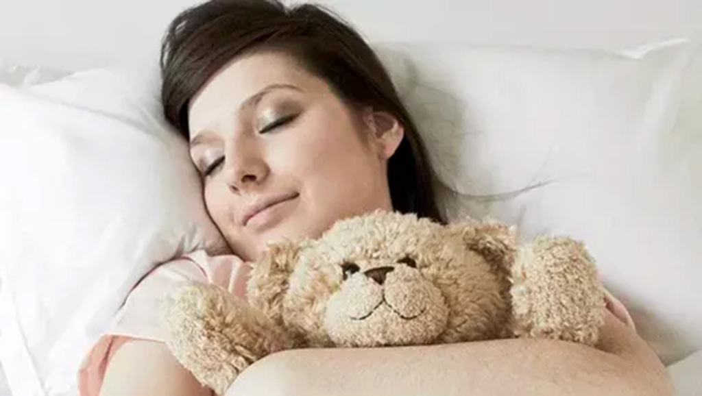 A woman peacefully sleeping on a white pillow while hugging a soft beige teddy bear close to her chest.