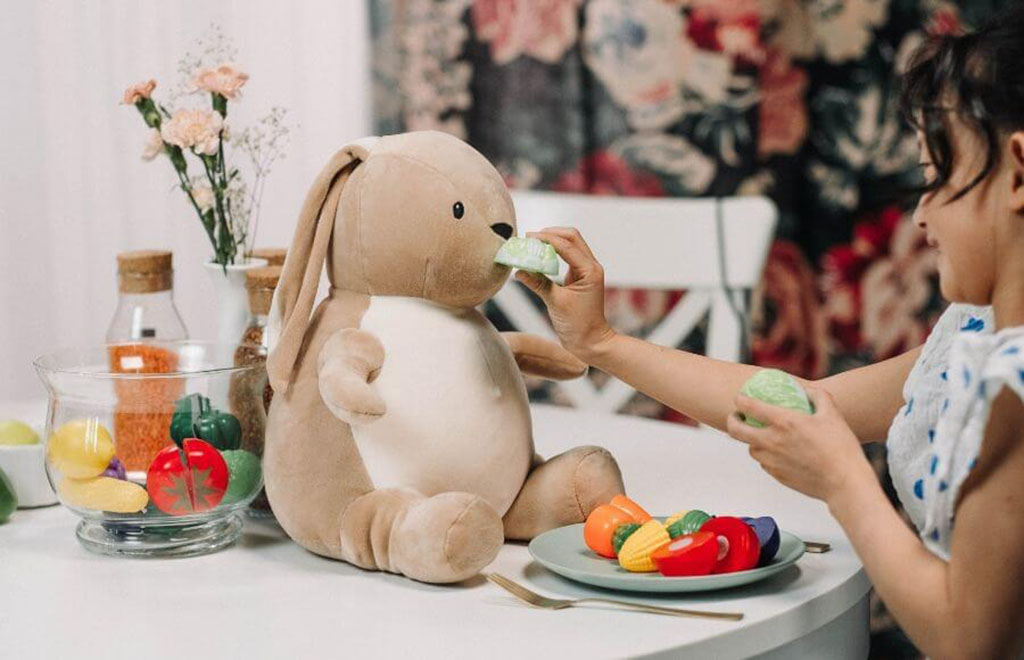 Little girl sitting at a table, playfully feeding a soft brown bunny plush toy with pretend vegetables during imaginative playtime.
