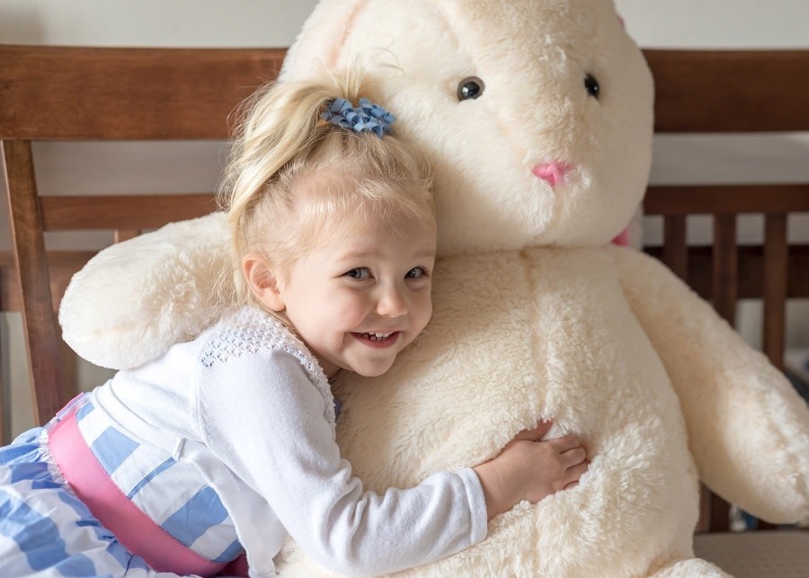 A smiling young girl hugs a large fluffy white bunny plush toy while sitting on a wooden bed.