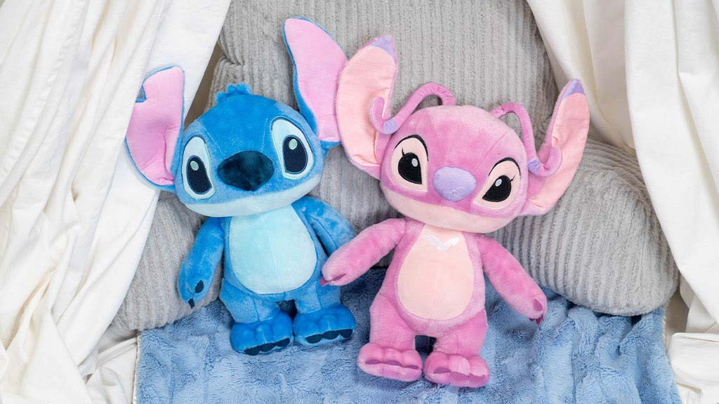 Two Stitch-themed plush toys, one blue and one pink, sitting together on a soft grey chair with a cozy blanket and white curtains around them.