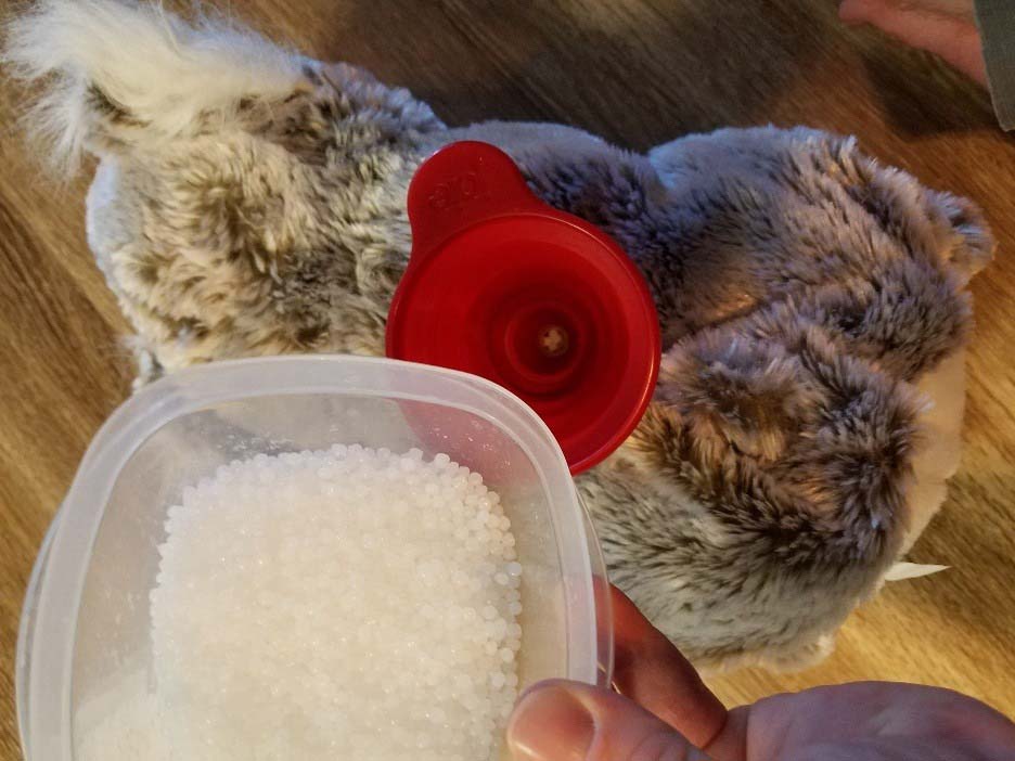 Hands filling a plush toy with white poly pellet beads using a red funnel for added weight and firmness.