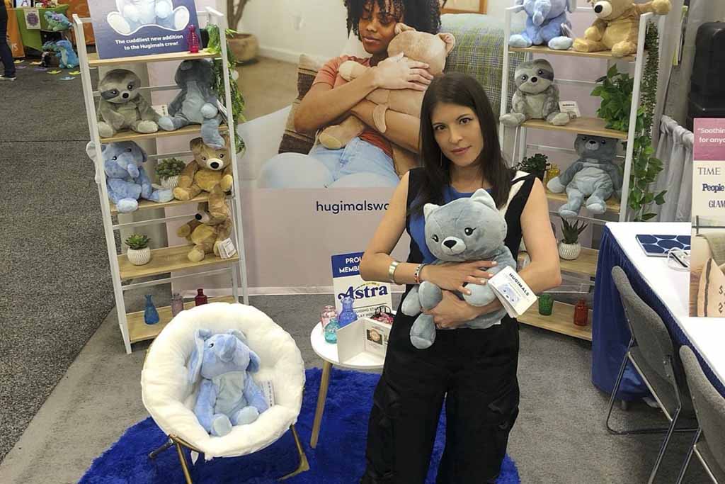 Woman holding a grey weighted plush bear at a trade show booth featuring shelves of Hugimals stuffed animals, display signage, and seating setup.
