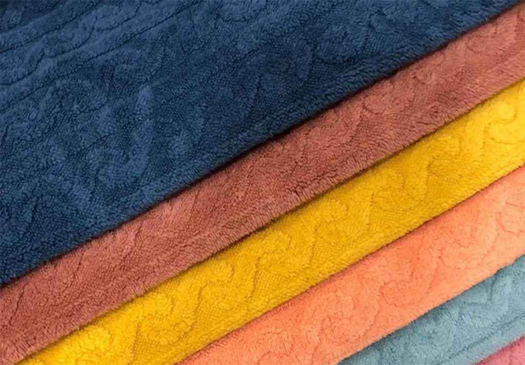 Stacks of textured plush fabric in blue, brown, yellow, orange, and pastel tones, showing material options commonly used for plush toys, stuffed animals, and soft baby products.