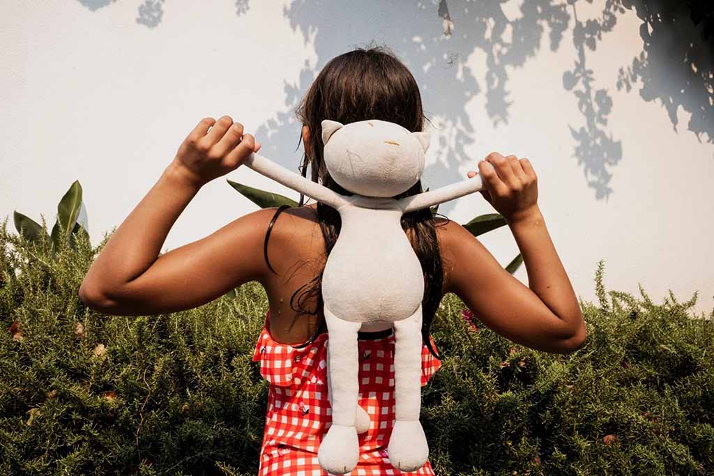  A child in a red checkered dress holding a long-limbed plush toy behind their neck while standing in front of greenery and a sunlit wall.