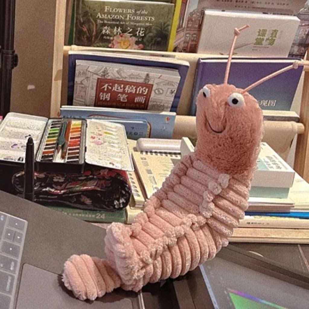 A pink caterpillar plush toy with a smiling face and bendable antennae sitting on a desk surrounded by books, a watercolor set, and stationery.