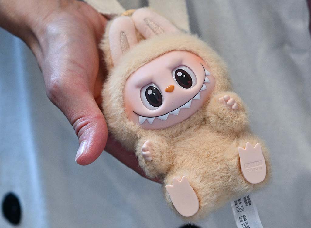 Person holding a small beige Labubu plush keychain with a wide grin, big eyes, and bunny ears, showing detailed textures and cute expression.