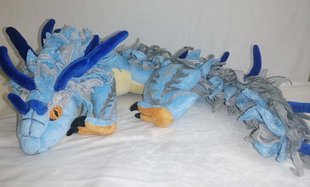A detailed blue and gray dragon plush toy with long body, embroidered eyes, soft fleece mane, and fabric horns, displayed on a white background.