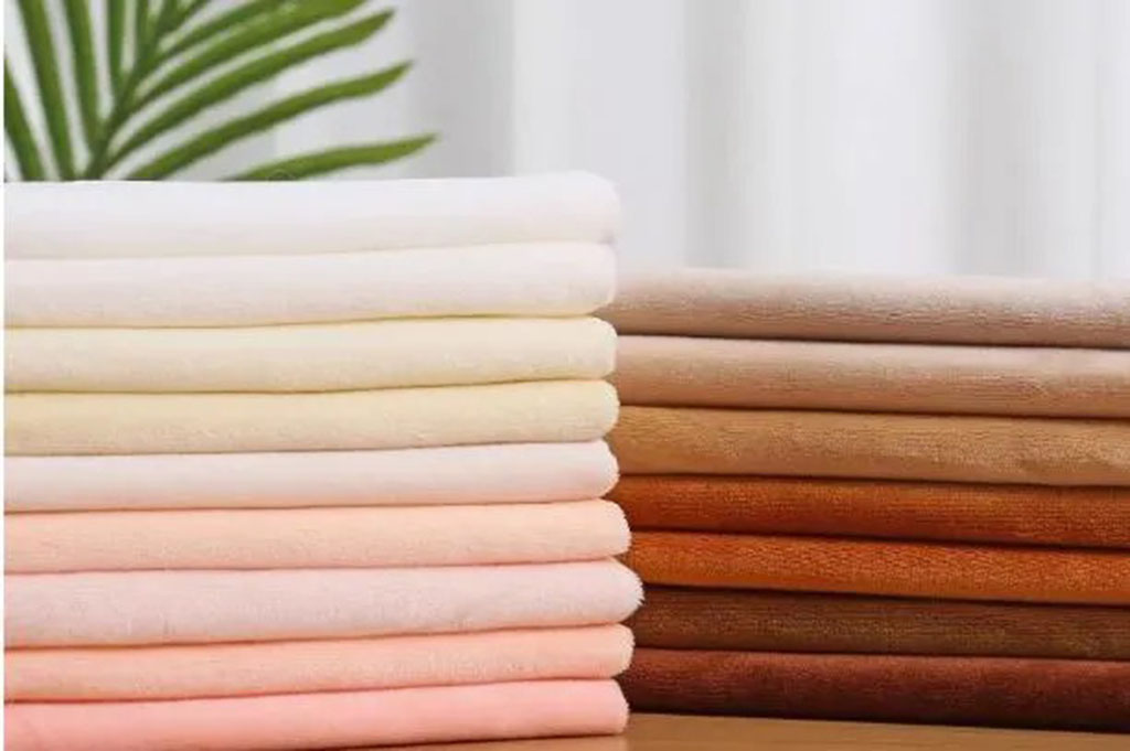 Stacked plush fabric sheets in soft skin-tone and pastel colors, neatly arranged in two columns on a table for plush toy manufacturing.
