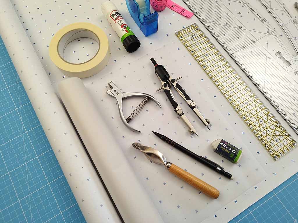 A flat lay of essential plush pattern-drafting tools including rulers, compass, tracing paper, masking tape, glue stick, measuring tape, punch tool, pencil, ergonomic awl, and drafting board on a blue cutting mat.