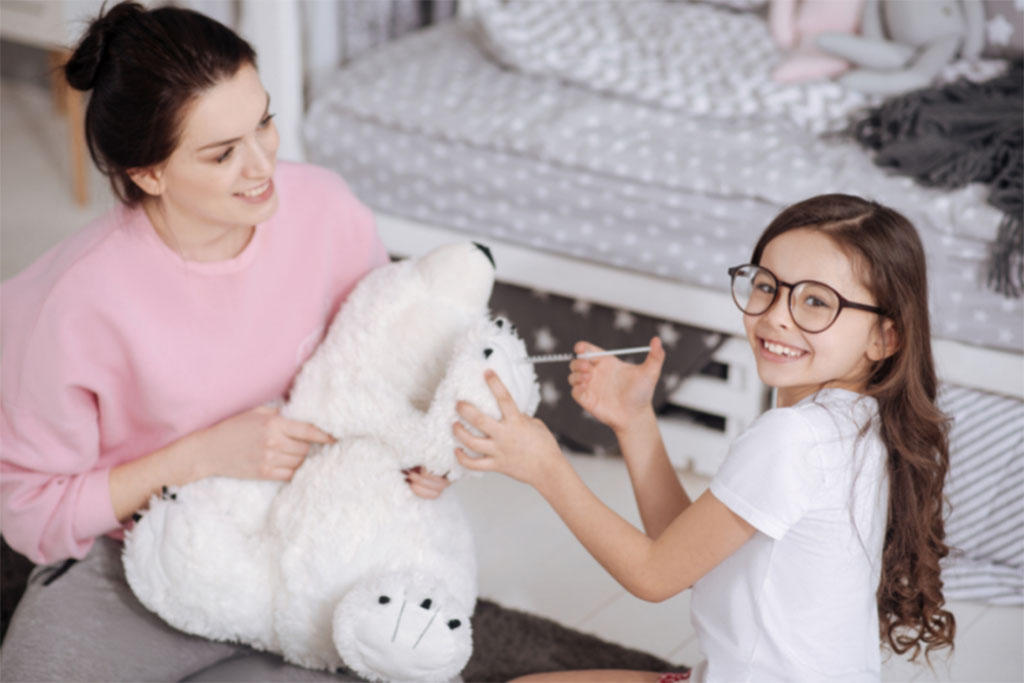Smiling mother and daughter sitting on the floor together, playfully brushing a large white polar bear plush toy in a cozy bedroom setting.