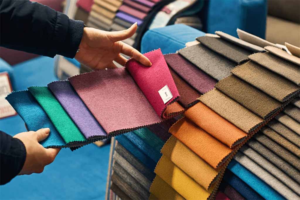 Person selecting from a range of colorful fabric swatches including blue, green, purple, red, and orange tones, displayed for texture and color comparison in material selection.