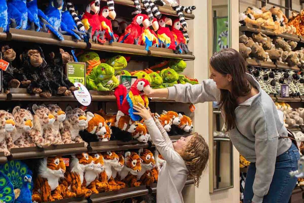 A child reaching for a colorful parrot plush toy with an adult’s help in a store filled with various animal plush toys on shelves.