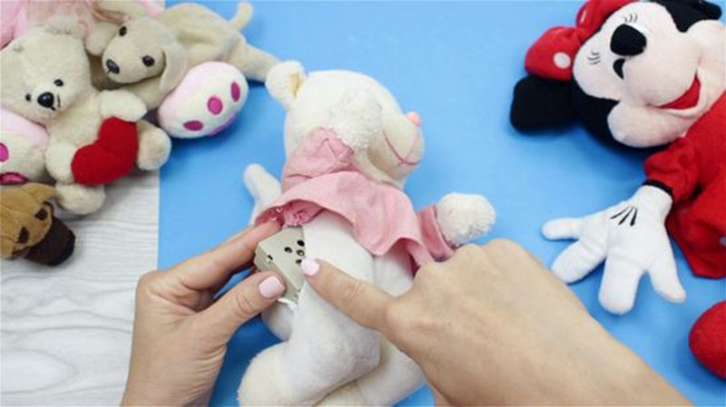 Hands cleaning a white stuffed teddy bear using a small metal brush while other plush toys sit nearby on a blue surface.