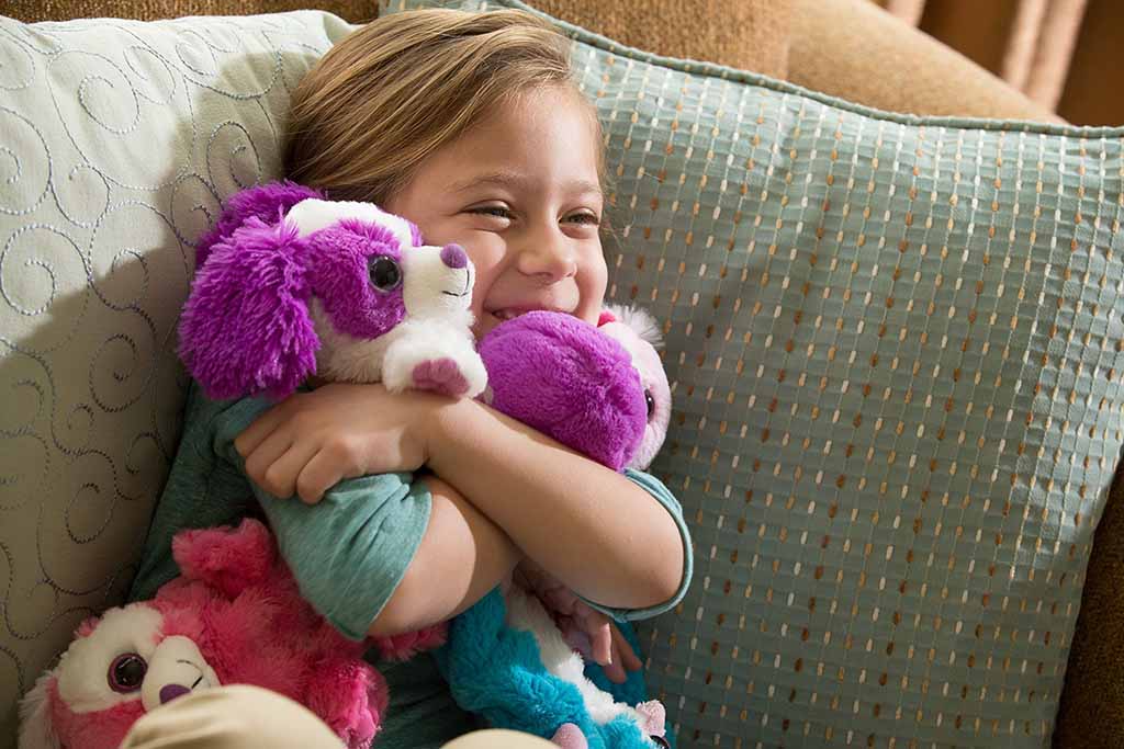A smiling young child sits on a couch hugging several bright purple and white plush puppy toys, looking happy and comfortable.