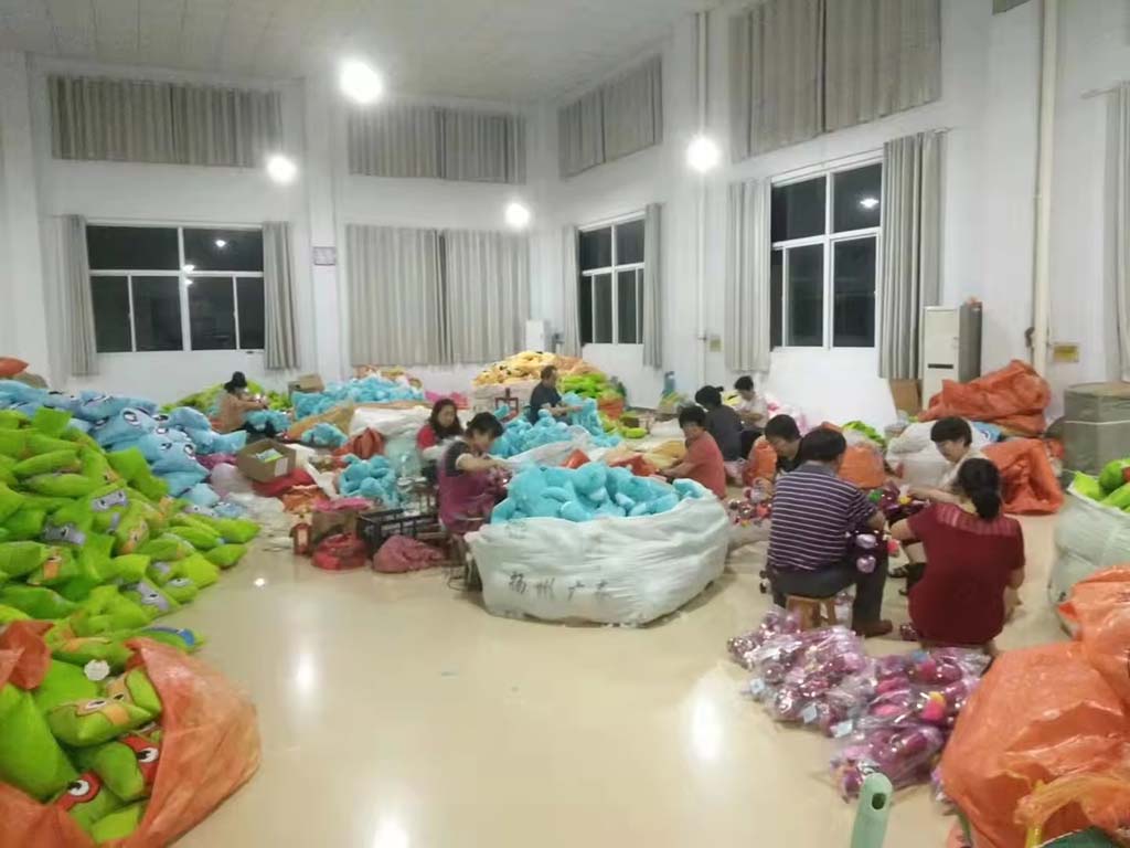 Workers in a plush toy factory sorting and packing large batches of stuffed animals, with piles of colorful plush toys and large storage bags spread across the production floor.