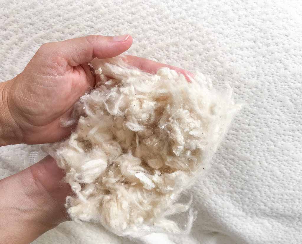 Hands holding a soft bundle of natural off-white kapok fiber stuffing over a textured white surface, showing its lightweight, fluffy texture.