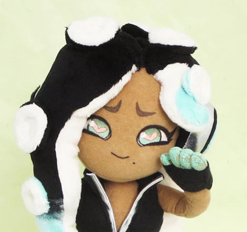 Close-up of a plush doll with embroidered heart-shaped eyes, brown skin tone, and black-and-white hair with aqua accents, raising one hand as if waving.