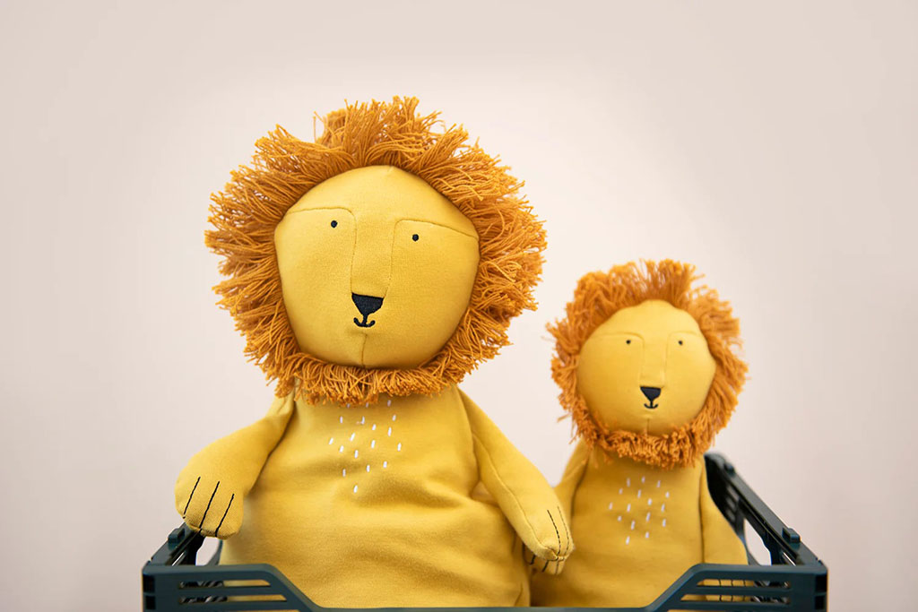 Two yellow lion plush toys with simple embroidered faces and yarn manes, placed in a crate against a neutral background, showcasing modern minimalist plush design.