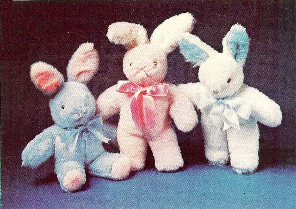 Three soft vintage-style bunny plush toys in blue, pink, and white, each with a ribbon bow, standing together against a dark background.