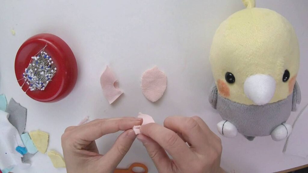 Hands sewing small pink plush fabric pieces beside a pin holder, fabric scraps, and a finished yellow-and-gray bird plush toy on a white crafting table.