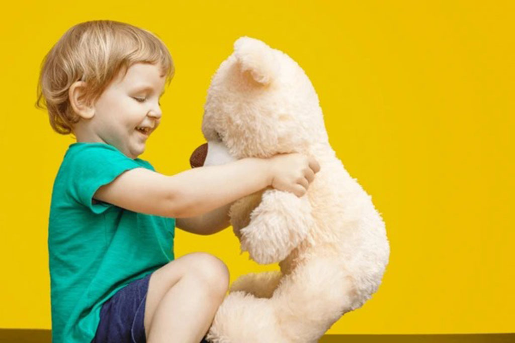 Smiling young boy in a turquoise shirt holding and hugging a soft white teddy bear against a bright yellow background, expressing warmth and joy.