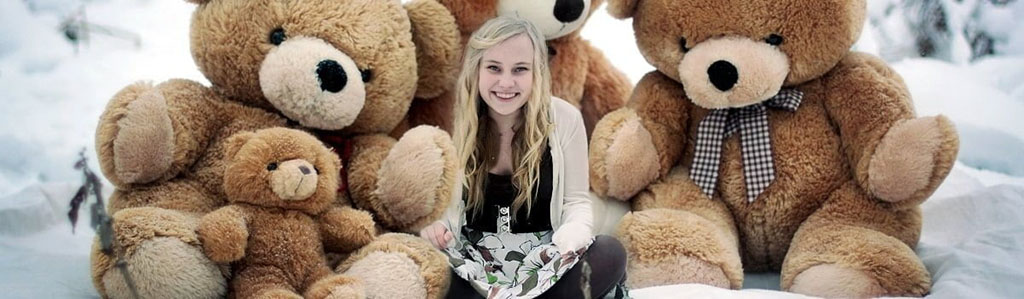A smiling girl sitting outdoors in the snow surrounded by large brown teddy bears of different sizes, creating a cozy winter scene.