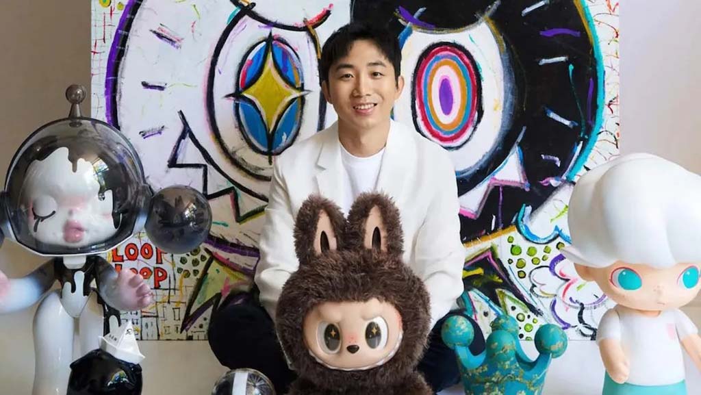 Artist sitting with Labubu and other Pop Mart art toys in front of a colorful abstract painting background.