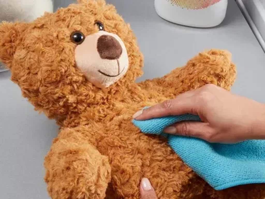 A person wiping the surface of a brown teddy bear with a blue cloth to remove dirt and stains.