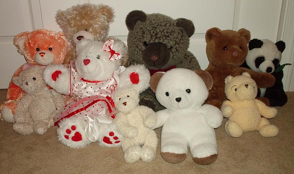 A group of assorted teddy bears in various colors, sizes, and styles sitting together on a carpeted floor, including classic brown bears, white bears, themed bears, and a panda plush.