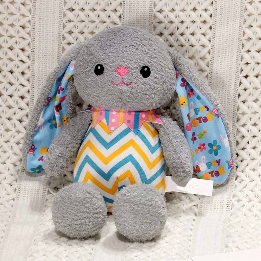 A soft gray bunny plush toy with colorful patterned ears and a zigzag outfit, sitting on a textured white surface.