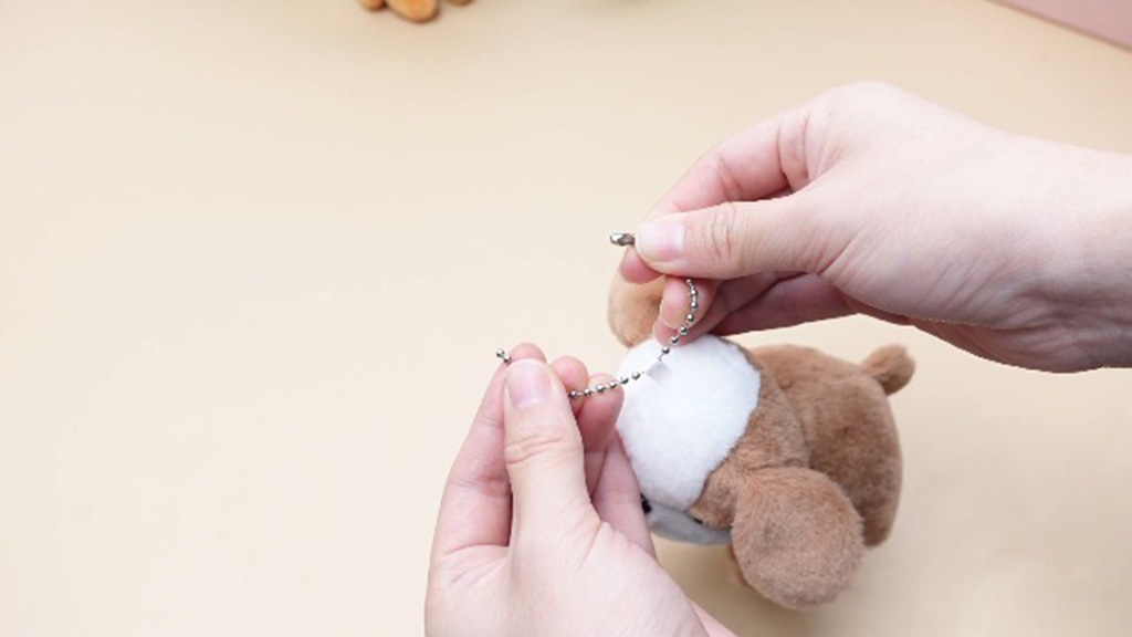 Hands connecting a small metal ball-chain to a brown plush toy to attach it as a keychain accessory.
