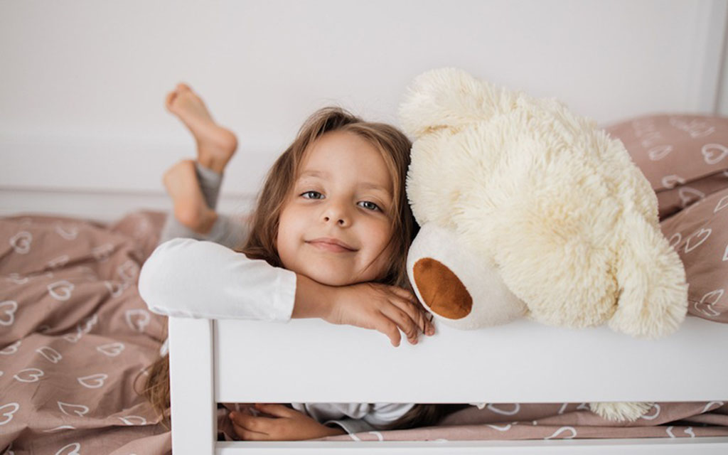 A smiling young girl lying in bed beside a large cream-colored teddy bear on soft heart-patterned bedding.