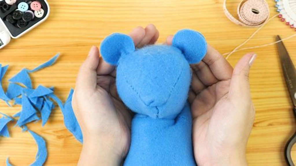 Hands holding a partially sewn blue teddy bear head on a wooden crafting table, surrounded by fabric scraps, buttons, thread, and scissors.