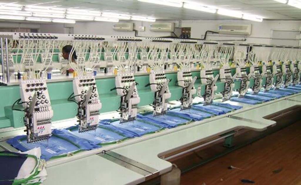 A row of automated multi-head embroidery machines simultaneously stitching designs onto multiple fabric pieces in a factory.