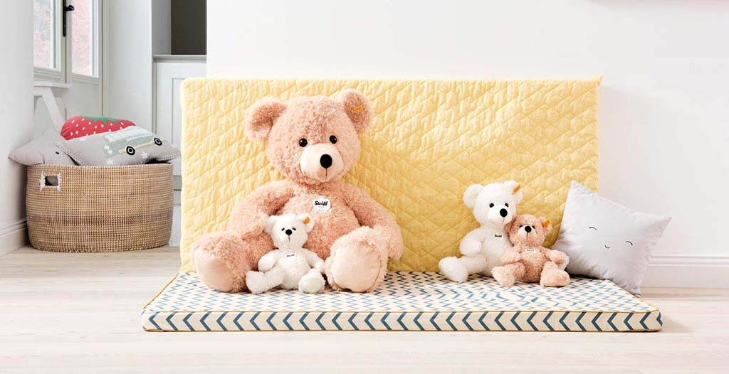A group of soft teddy bears of different sizes arranged on a patterned mat with yellow quilted backing and decorative pillows in a bright, cozy room.