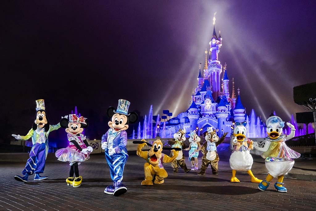 Disney characters including Mickey, Minnie, Goofy, Donald, and others posing in front of the illuminated Sleeping Beauty Castle at Disneyland Paris.