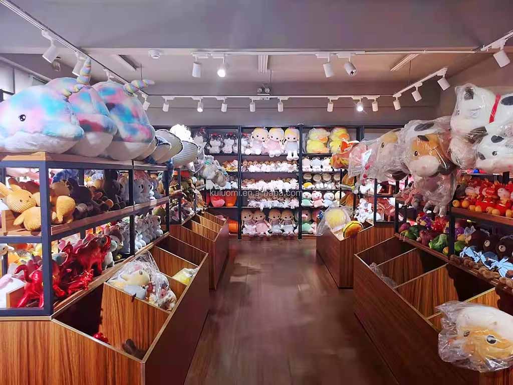 A brightly lit retail store filled with shelves of colorful plush toys arranged in rows, including large stuffed animals on the top shelves and smaller plushies in wooden bins along the aisle.
