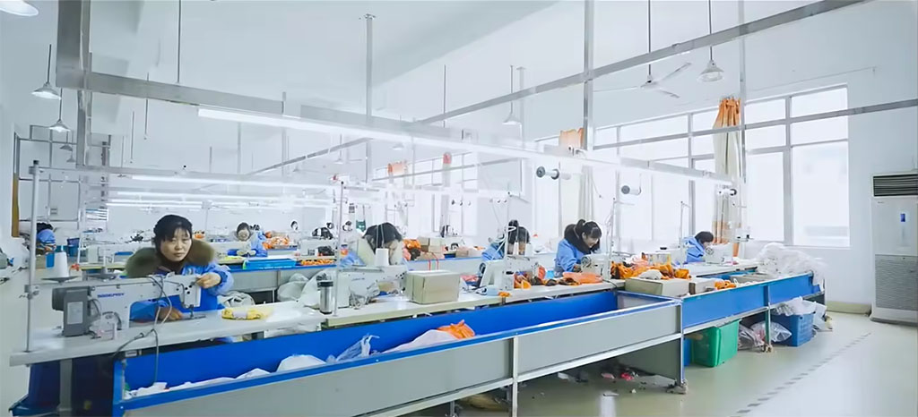Workers in a bright, organized factory sewing plush toys at multiple workstations, with sewing machines, materials, and tools arranged along long production tables.