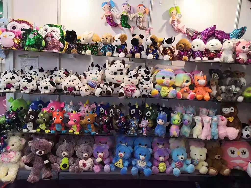 A large wall display filled with colorful plush toys, including sequin animals, cows, dragons, bears, unicorns, and mermaid dolls, arranged neatly on multi-level shelves at a toy exhibition.