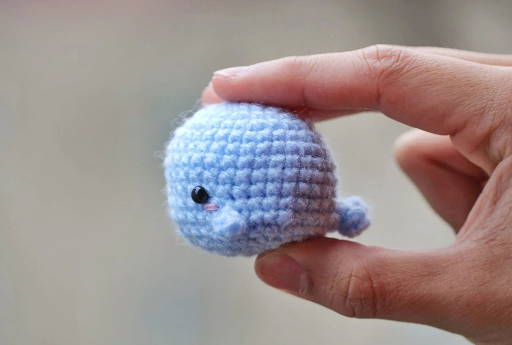 Hand holding a small blue crocheted whale plush, showing close-up stitch details and cute embroidered face, representing handmade amigurumi-style soft toys.