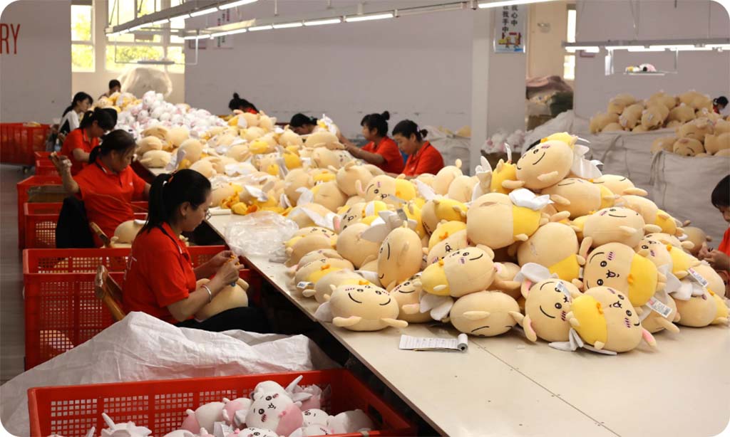 Factory workers in red uniforms assembling plush toys at long production tables, with large piles of finished soft dolls and red sorting baskets arranged throughout the workspace.